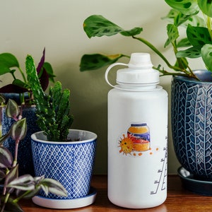 May include: A white water bottle with a colorful sticker of a jar of orange marmalade and a sun. The bottle has a white lid and a measurement scale on the side. The bottle is sitting on a wooden surface next to three potted plants.