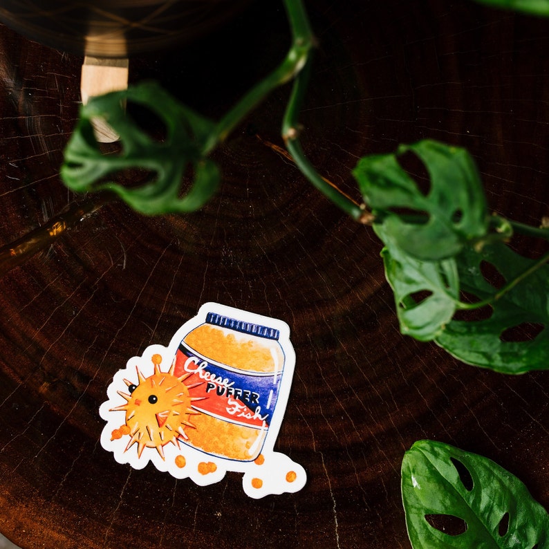 May include: A white sticker with a cartoon illustration of a jar of "Cheese Puffer Fish" with a puffer fish next to it. The jar is orange and blue with white lettering. The puffer fish is yellow with black eyes and spines.