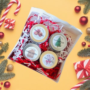 May include: A white gift box filled with red and white shredded paper, containing four round tins with labels. The labels read "Caramel Apple", "Christmas Morning", "Hansel & Gretel", and "Candy Cane". Christmas ornaments and candy canes surround the box.