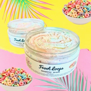 Op de afbeelding: Twee potten met geklopt zeep met een kleurrijk swirl-ontwerp. De potten zijn gelabeld "Froot Loops Whippin' Wash" en hebben een kleurrijk ontbijtgraanontwerp op het etiket. De potten staan op een roze en gele achtergrond.