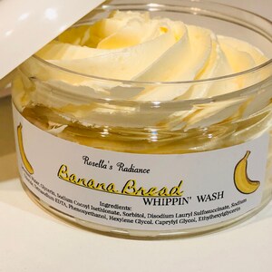 May include: A clear jar with a white lid, filled with a light yellow, whipped product. The jar has a label with the text "Banana Bread WHIPPIN' WASH" and a banana illustration. The product is from "Rosella's Radiance."