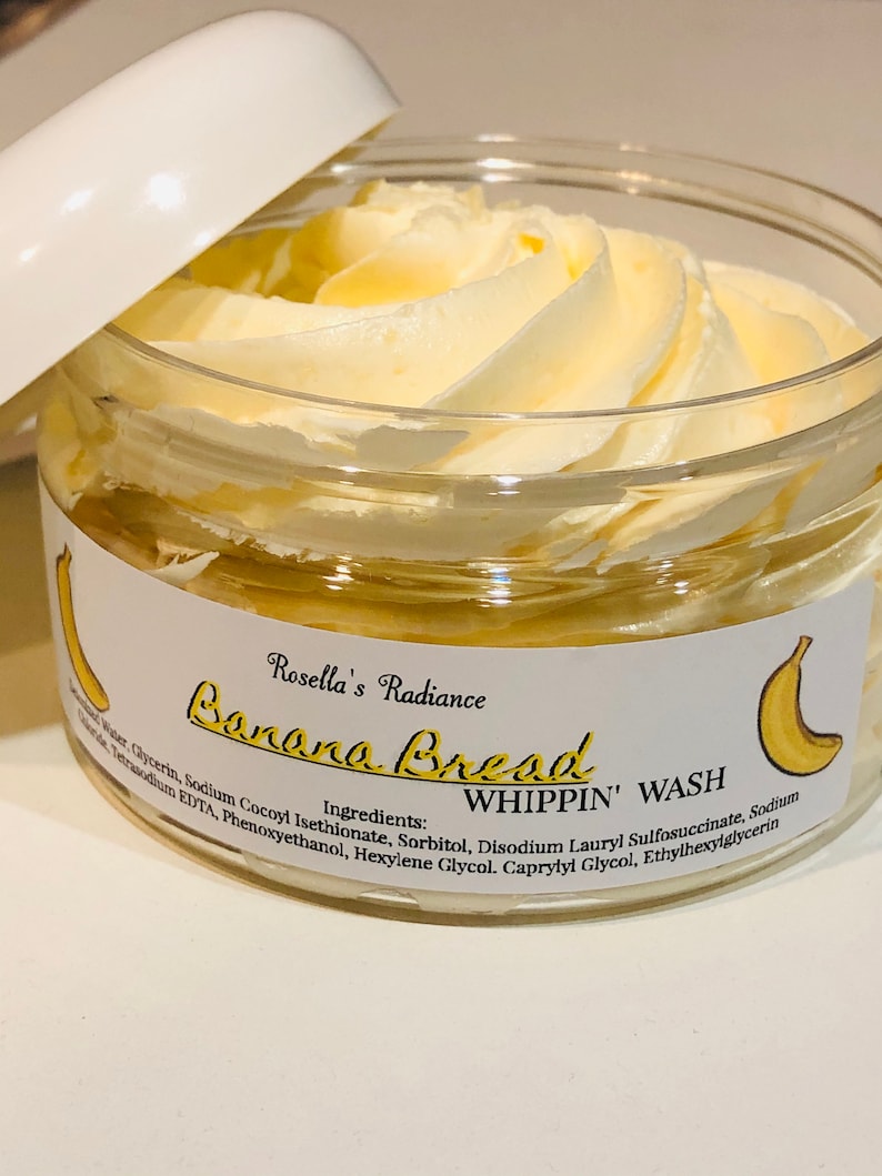 May include: A clear jar with a white lid, filled with a light yellow, whipped product. The jar has a label with the text "Banana Bread WHIPPIN' WASH" and a banana illustration. The product is from "Rosella's Radiance."