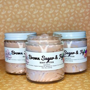 Puede incluir: Tres frascos de vidrio de manteca corporal con tapas blancas. Los frascos est&aacute;n etiquetados como "Brown Sugar & Fig Body Butter" y contienen una manteca corporal batida de color marr&oacute;n claro. Los frascos est&aacute;n sobre un fondo marr&oacute;n claro.