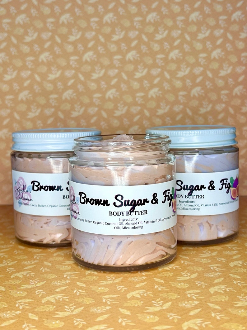 Puede incluir: Tres frascos de vidrio de manteca corporal con tapas blancas. Los frascos est&aacute;n etiquetados como "Brown Sugar & Fig Body Butter" y contienen una manteca corporal batida de color marr&oacute;n claro. Los frascos est&aacute;n sobre un fondo marr&oacute;n claro.