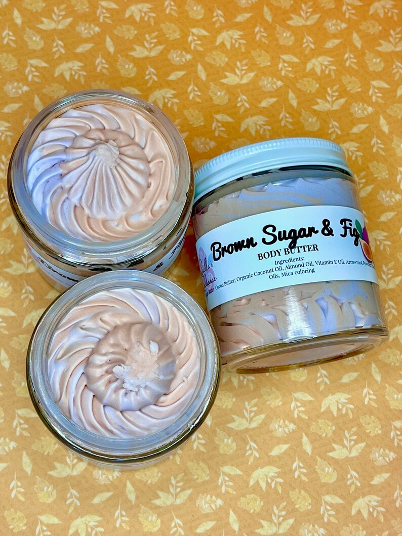 Puede incluir: Dos tarros de vidrio de manteca corporal con un remolino de crema p&uacute;rpura y beige. El tarro en primer plano est&aacute; etiquetado como "Brown Sugar & Fig Body Butter" con los ingredientes enumerados debajo.