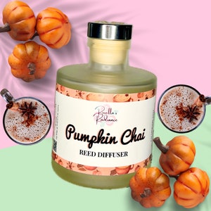 Może przedstawiać: Przezroczysta szklana butelka ze złotą nakrętką i etykietą z napisem "Pumpkin Chai Reed Diffuser" z jesiennym wzorem. Butelka jest otoczona małymi pomarańczowymi dyniami i dwoma kubkami chai.