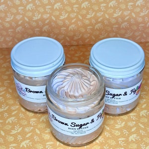 Puede incluir: Tres frascos de vidrio con tapas blancas llenos de manteca corporal marr&oacute;n claro. Los frascos est&aacute;n etiquetados como "Brown Sugar & Fig Body Butter" y enumeran los ingredientes: Manteca de coco, aceite de coco org&aacute;nico, aceite de almendras, aceite de vitamina E y colorante mica.