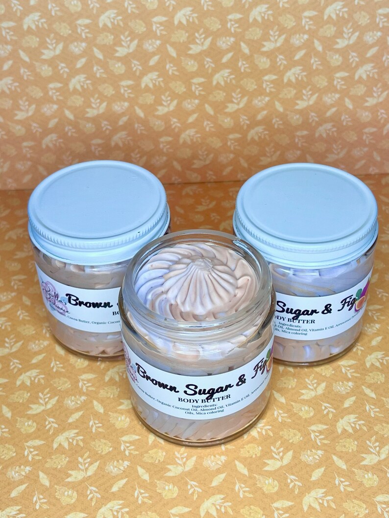 Puede incluir: Tres frascos de vidrio con tapas blancas llenos de manteca corporal marr&oacute;n claro. Los frascos est&aacute;n etiquetados como "Brown Sugar & Fig Body Butter" y enumeran los ingredientes: Manteca de coco, aceite de coco org&aacute;nico, aceite de almendras, aceite de vitamina E y colorante mica.