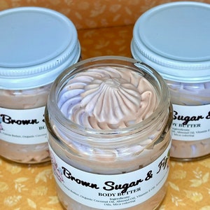 Puede incluir: Tres frascos de vidrio con tapas blancas llenos de manteca corporal marr&oacute;n claro y p&uacute;rpura. Los frascos est&aacute;n etiquetados como "Brown Sugar & Fig" y "Body Butter". Los ingredientes se enumeran como: Manteca de cacao, aceite de coco org&aacute;nico, aceite de almendras, aceite de vitamina E, colorante mica.