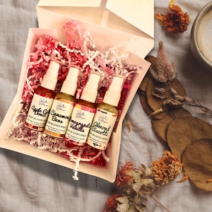 May include: A set of four small glass bottles with spray tops in a white gift box. The bottles are filled with a clear liquid and have labels with different scents: Apple Cider Donut, Cinnamon Buns, Pomegranate Vanilla, and Almond Biscotti.