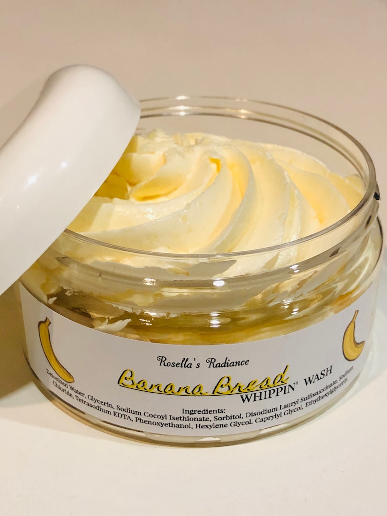 May include: A glass jar containing a light yellow, whipped cream-like product. The jar has a white label with the text "Rosella's Radiance Banana Bread WHIPPIN' WASH" and a banana illustration. The lid is white and partially open.