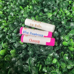 Puede incluir: Tres tubos de bálsamo labial con diferentes sabores: Vainilla, Frambuesa Azul y Cereza. El tubo de Vainilla es blanco con texto dorado, el tubo de Frambuesa Azul es rosa con texto azul y el tubo de Cereza es rosa con texto rojo. Los bálsamos labiales están sobre un fondo verde frondoso.