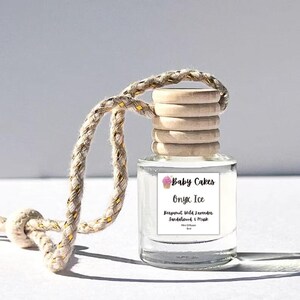 May include: A clear glass bottle with a wooden cap and a woven rope hanger. The bottle has a label that reads "Baby Cakes Onyx Ice" with scent details. The rope is beige with gold accents. The product is a car air freshener.