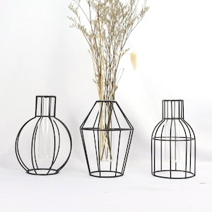 Könnte beinhalten: Drei schwarze Metallvasen mit Glasröhrchen im Inneren. Die Vasen haben unterschiedliche Formen: rund, Diamant und rechteckig. Die Vasen stehen auf einer weißen Oberfläche.