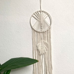 Może przedstawiać: Biały łapacz snów z makramą o okrągłym wzorze i pojedynczym piórem zwisającym z dołu.
