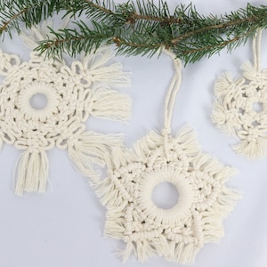 May include: Three white crocheted snowflake ornaments with fringe. The snowflakes are hanging from a branch of a pine tree.