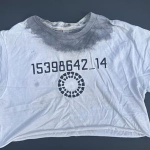 May include: White, cropped t-shirt with a crew neck and short sleeves. The shirt features the number sequence "15398642_14" printed in black, along with a circular design. The collar has a gray stain.