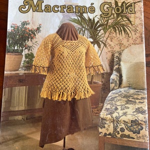 May include: A book titled "Macrame Gold" featuring a crocheted top with fringe on a dress form. The top is yellow and the dress form is wearing a brown skirt. The book is set in a room with a floral patterned wallpaper and a chair with a floral patterned cushion.