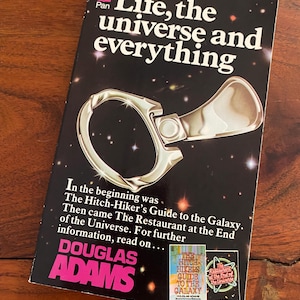 May include: A paperback book cover with a black background and a silver pull tab. The title is "Life, the universe and everything" by Douglas Adams. The book cover also features the text "The Hitch-Hiker's Guide to the Galaxy" and "The Restaurant at the End of the Universe."