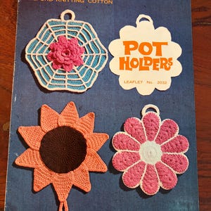 May include: A vintage Strutt's Milford Knitting Cotton leaflet featuring three crocheted pot holders. Designs include a white web pattern with a pink flower, a peach sunflower with a dark brown centre, and a pink and white daisy-shaped holder.