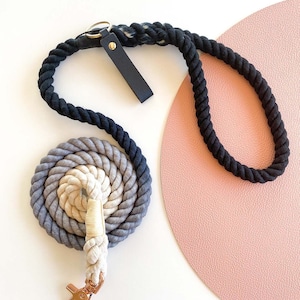 Puede incluir: Una correa de perro de cuerda negra y gris con un mango de cuero negro y un clip de metal dorado. La correa está enrollada sobre una superficie rosa.