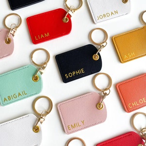 May include: A collection of personalised leather keychains with gold hardware and various names, including Liam, Jordan, Ash, Sophie, Abigail, Emily, Chloe. The keychains are displayed on a white surface.