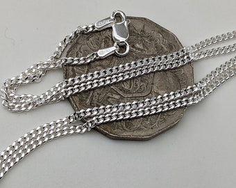 Könnte beinhalten: Eine silberne Curb-Kette mit einem Karabinerverschluss. Die Kette ist in der Mitte gefaltet und auf einer Silbermünze platziert.