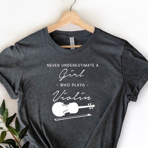 May include: Dark gray t-shirt with white text that reads "Never underestimate a girl who plays violin" and a white illustration of a violin with a bow.