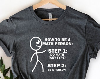 May include: A dark gray t-shirt with a white stick figure and text that reads "HOW TO BE A MATH PERSON: STEP 1: DO MATH (ANY TYPE) STEP 2: BE A PERSON"