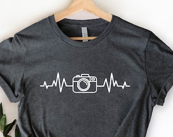 May include: A dark grey t-shirt with a white camera graphic and a heartbeat line. The shirt is on a hanger and has a white background.