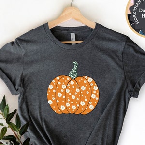 Puede incluir: Una camiseta gris oscuro jaspeada con un diseño de calabaza floral. La calabaza es naranja con flores blancas y un tallo verde.