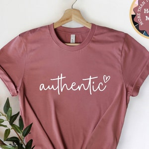 Puede incluir: Una camiseta de color malva jaspeado con la palabra "auténtico" escrita en cursiva blanca con un pequeño símbolo de corazón. La camiseta está colgada de una percha de madera sobre un fondo blanco.