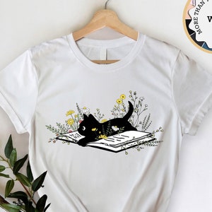May include: White t-shirt with a black cat reading a book surrounded by yellow flowers. The cat is looking at the book. The text "MORE THAN 14 COLORS AND SIZES" is in a circle above the cat.
