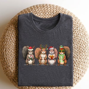 Puede incluir: Una camiseta gris oscuro con un gráfico de cuatro ardillas con gorros y accesorios navideños. Las ardillas sostienen bastones de caramelo, adornos y un regalo.