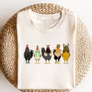 May include: A white t-shirt with a graphic of five chickens wearing Christmas hats and lights. The chickens are in a row, facing forward. The background is white.