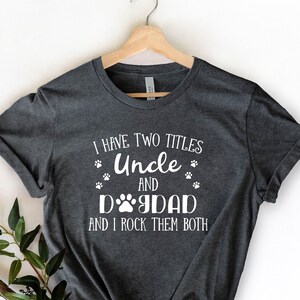 Puede incluir: Una camiseta gris oscuro con texto blanco que dice "Tengo dos títulos Tío y Papá Perro y los rockeo ambos". La camiseta tiene un gráfico de huella de pata.