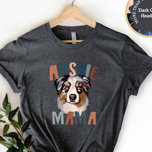 May include: A dark grey t-shirt with a colourful "Aussie Mama" graphic featuring a cartoon Australian Shepherd dog. The shirt is on a hanger with a small sign that says "More than 14 colours and sizes".
