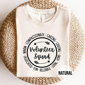 May include: A natural-colored t-shirt with the words "Volunteer Squad" in a black circular design. The design includes words like "compassionate," "caring," and "loyal." The word "NATURAL" is printed in a white rectangle.