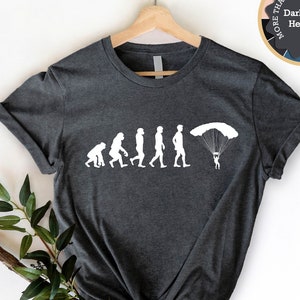 May include: A dark heather grey t-shirt with a white graphic of the evolution of man, ending with a man skydiving with a parachute. The text "More Than 14 Colors and Sizes" is on a circle with a rainbow of colors around the edge.