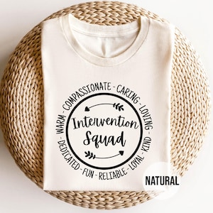 May include: A cream-colored t-shirt with the words "Intervention Squad" in a black circular design. The design includes words like "compassionate," "caring," and "loving." The word "NATURAL" is printed below the shirt.