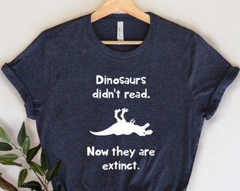 May include: A dark blue t-shirt with a white graphic of a dinosaur lying on its back. The text on the shirt reads "Dinosaurs didn't read. Now they are extinct."
