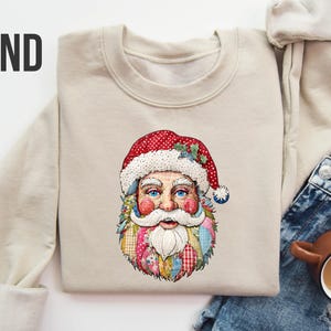 Puede incluir: Una sudadera de color arena con un gráfico colorido de Papá Noel. La ilustración de Papá Noel tiene un gorro rojo con lunares blancos, ojos azules y una larga barba blanca. La palabra "SAND" está impresa en letras negras grandes en la parte superior izquierda.
