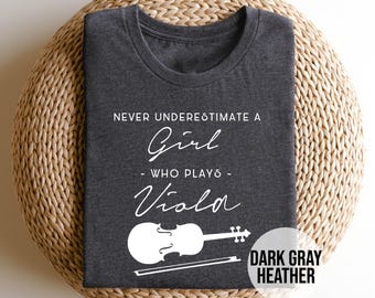 Puede incluir: Camiseta gris oscuro jaspeado con el texto blanco "NUNCA SUBESTIMES A UNA CHICA QUE TOCA LA VIOLA". Un gráfico blanco de viola y arco está debajo. La camiseta está doblada sobre una superficie tejida.