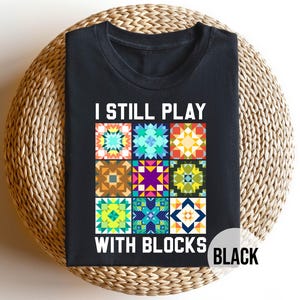 May include: Black t-shirt with the text "I STILL PLAY WITH BLOCKS" above a colorful quilt block design. The design features nine different quilt block patterns in various colors. The word "BLACK" is in a white circle at the bottom right.