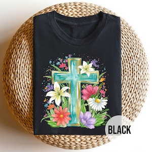 Puede incluir: Camiseta negra con una cruz turquesa con ribete dorado, rodeada de flores coloridas. El diseño incluye lirios blancos, margaritas rojas y rosas, y flores moradas. La palabra "BLACK" está impresa en la parte inferior derecha.