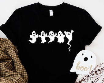 May include: Black t-shirt featuring a row of five white cartoon ghosts in various poses. A wooden ghost-shaped decoration with the word "boo" is visible. The shirt is folded, and a plaid fabric and jeans are in the background.