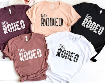 May include: Five t-shirts with the text "Let's Rodeo" printed on them in a white outline with black fill. The shirts are in various colors, including maroon, peach, white, and black.
