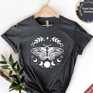 Puede incluir: Camiseta gris oscuro jaspeada con un gráfico blanco de una polilla con fases lunares y detalles florales. El texto "Más de 14 colores y tallas" es visible en un círculo con muestras de colores.