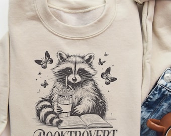 May include: A beige sweatshirt with a graphic of a raccoon holding a cup of coffee and a book. The raccoon is surrounded by butterflies. The text "BOOKTROVERT" is printed below the raccoon.