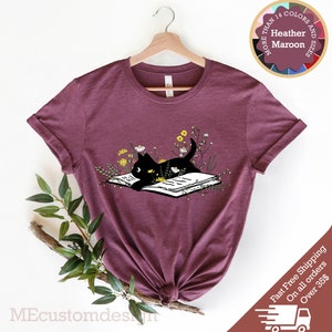 May include: A heather maroon t-shirt with a black cat reading a book surrounded by flowers. The text "More Than 14 Colors and Sizes" is on a circle with a variety of colored circles around it. The text "Heather Maroon" is in the center of the circle.